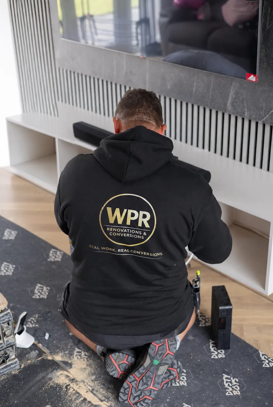 Shane Williams seen from behind, kneeling at a white media wall unit, wearing a black hoodie printed with the WPR Renovations and Conversions logo and the line Real work, real conversions